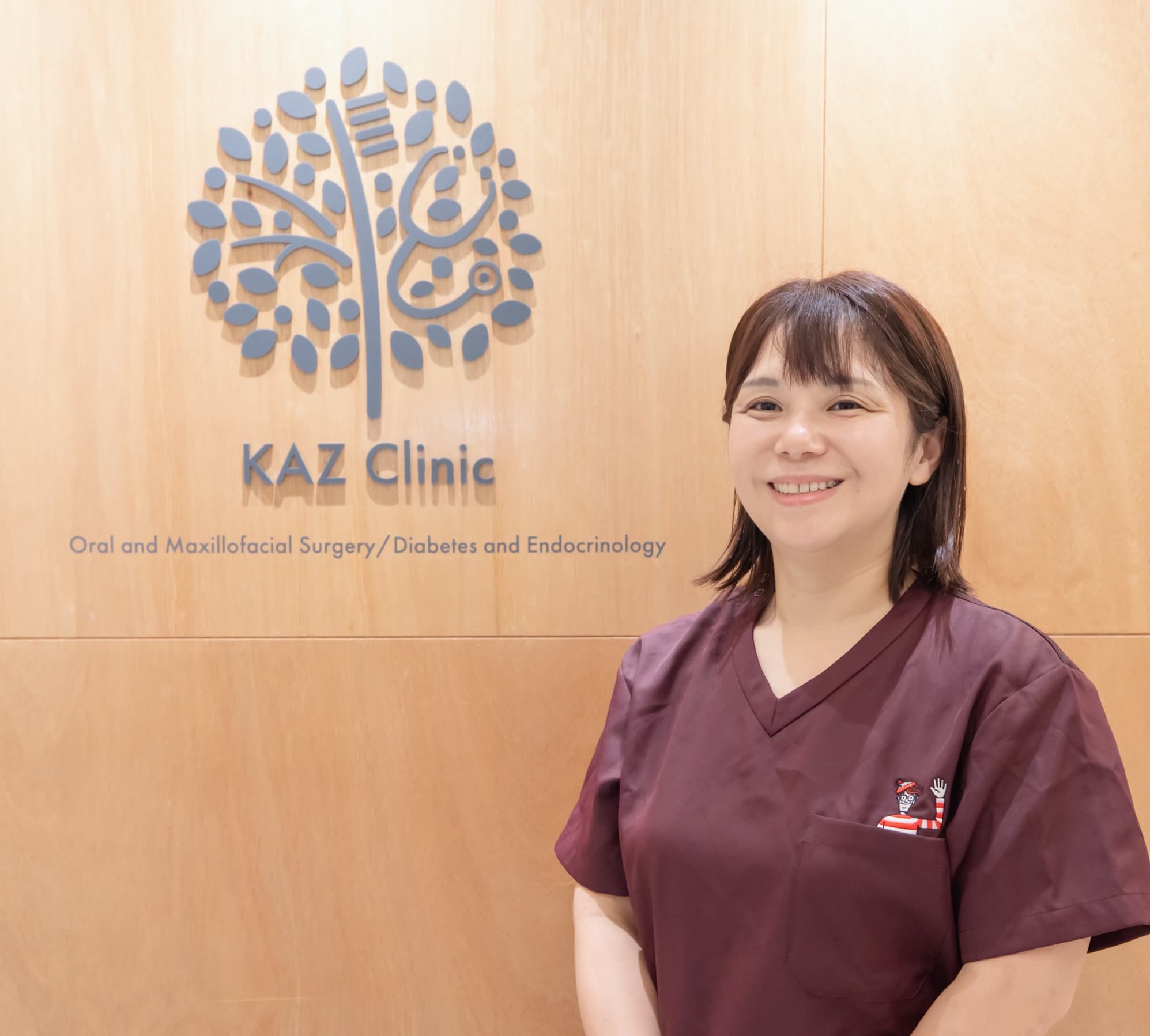 「KAZ Clinic」のロゴを背に笑顔で立つ、えんじ色のスクラブを着た女性医師