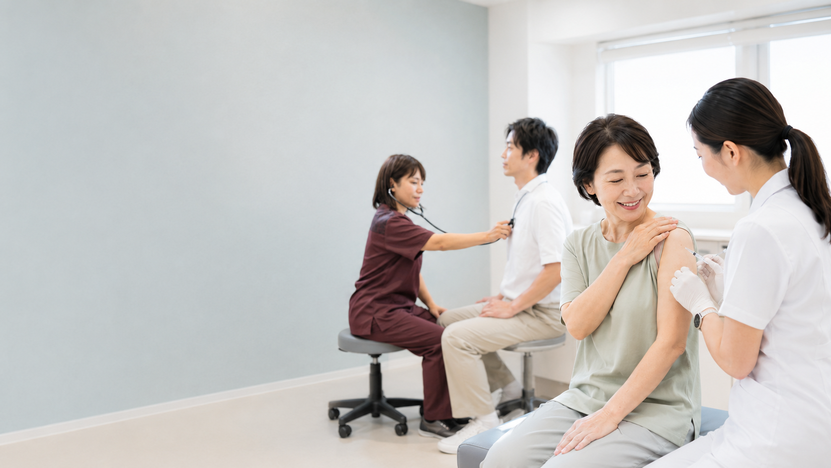 診察室で女性患者が予防接種を受け、奥では医師が男性患者を聴診する診療の様子